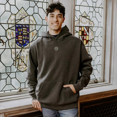 College-aged man wearing the Chi Phi embroidered black hoodie, standing in front of stained glass windows with fraternity-style crest designs.
