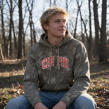 College-aged male wearing the fraternity Deadwood Camo Hoodie with red and white Chi Phi lettering, seated outdoors in a wooded setting.