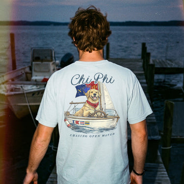 Young man standing on a dock at sunset wearing a light blue t-shirt featuring a golden retriever steering a sailboat with a flag and the phrase “Chasing Open Water” on the back.