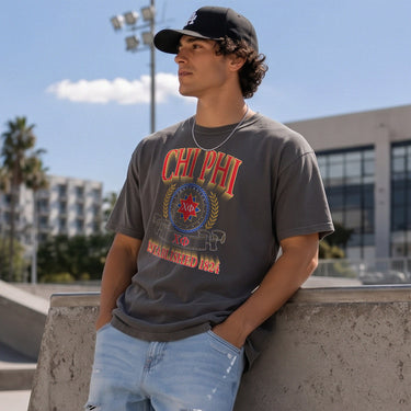 College-aged male wearing the fraternity Collegiate Crest Graphic Tee in charcoal grey, featuring vintage Chi Phi crest artwork on campus.